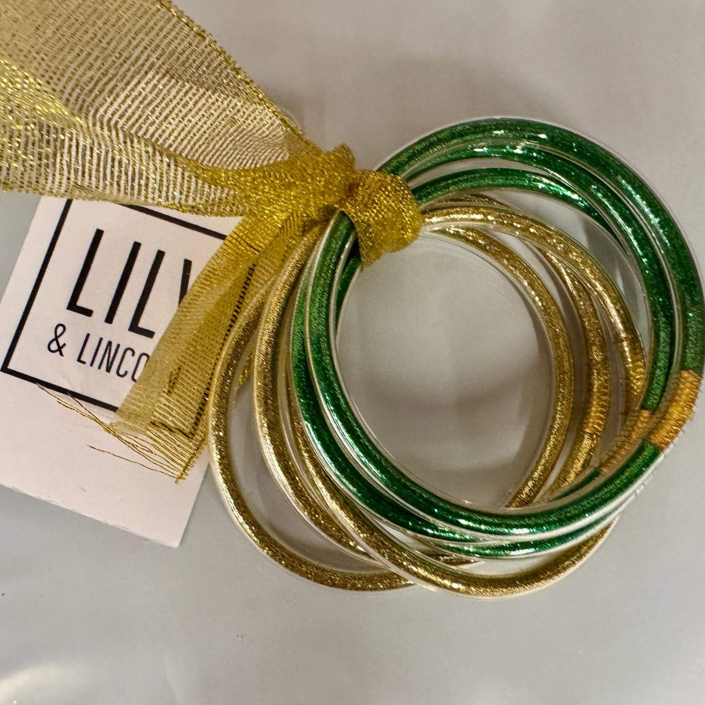 Set of green and gold bangles with a gold ribbon and 'LILY & LINCO' tag on a white background