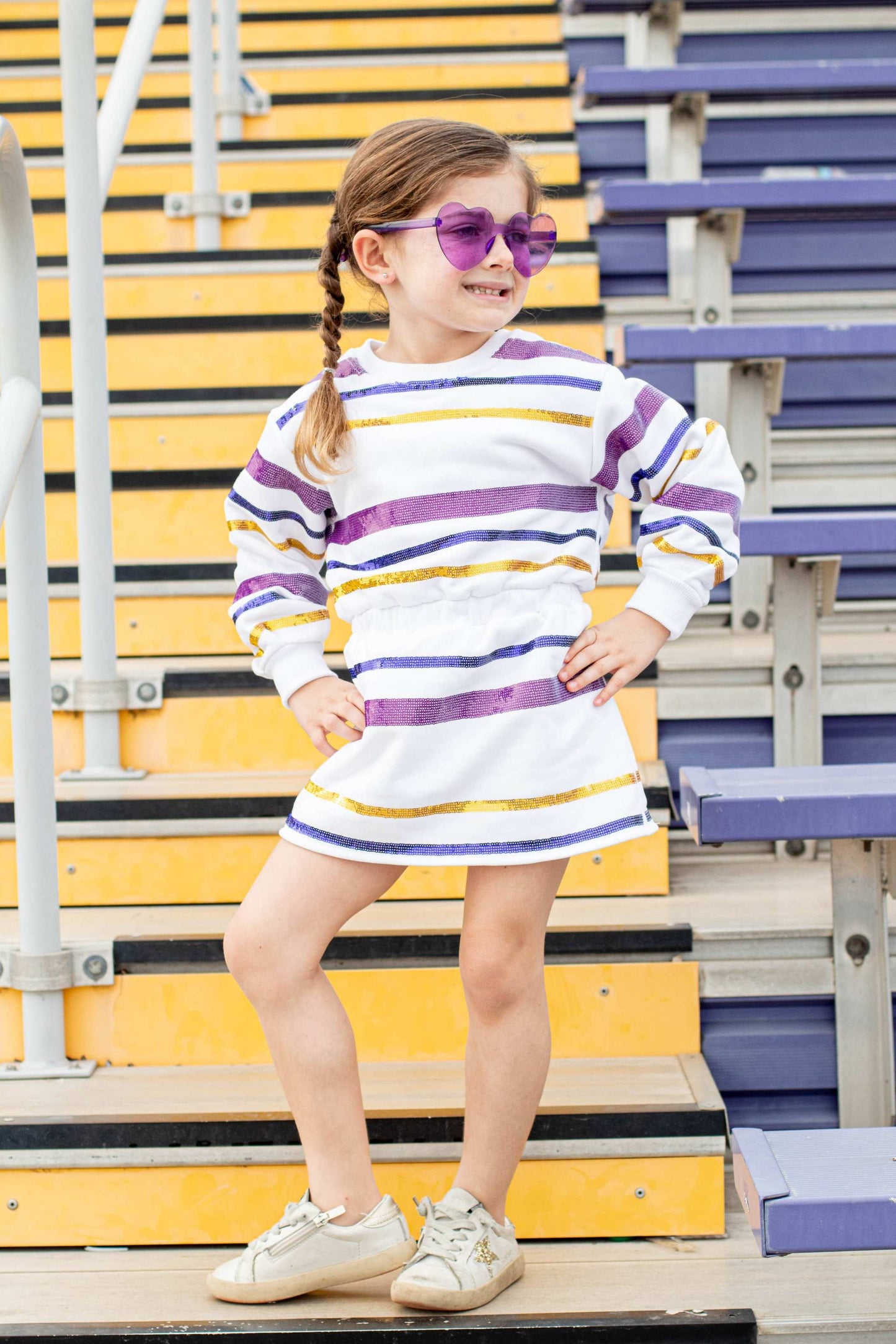 Girl wearing SeersuckerJOEY LLC Throw me the ball game day dress in purple and gold stripes, standing on bleachers.