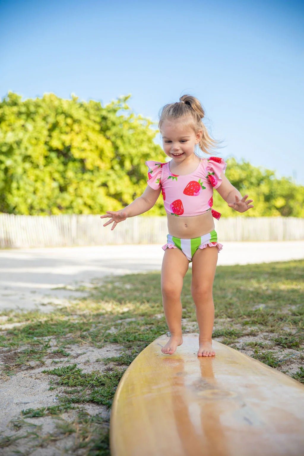 Toddler in Blueberry Bay Sweet Charlie strawberry swimsuit standing on a surfboard.