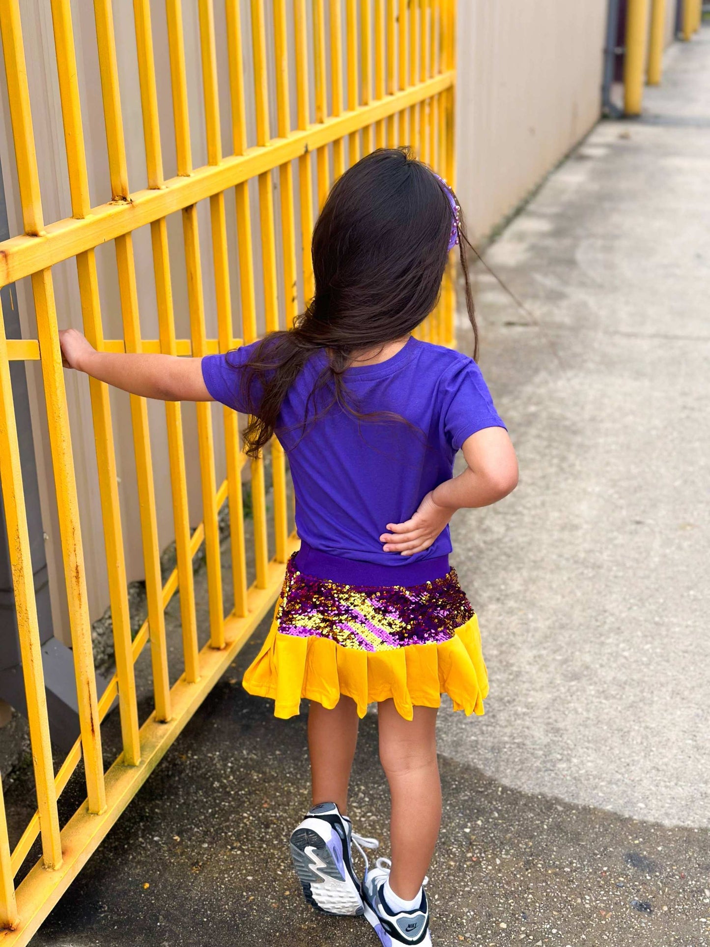 Child in purple shirt and sequined purple and gold skort near a yellow fence, showcasing Mardi Gras Creations.