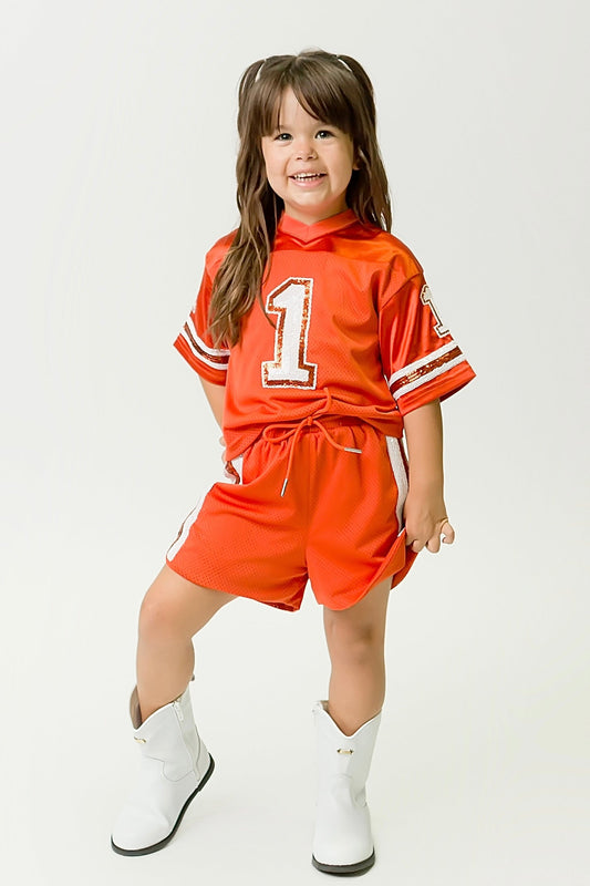 Child wearing burnt orange and white sequin jersey with number 1 and matching shorts
