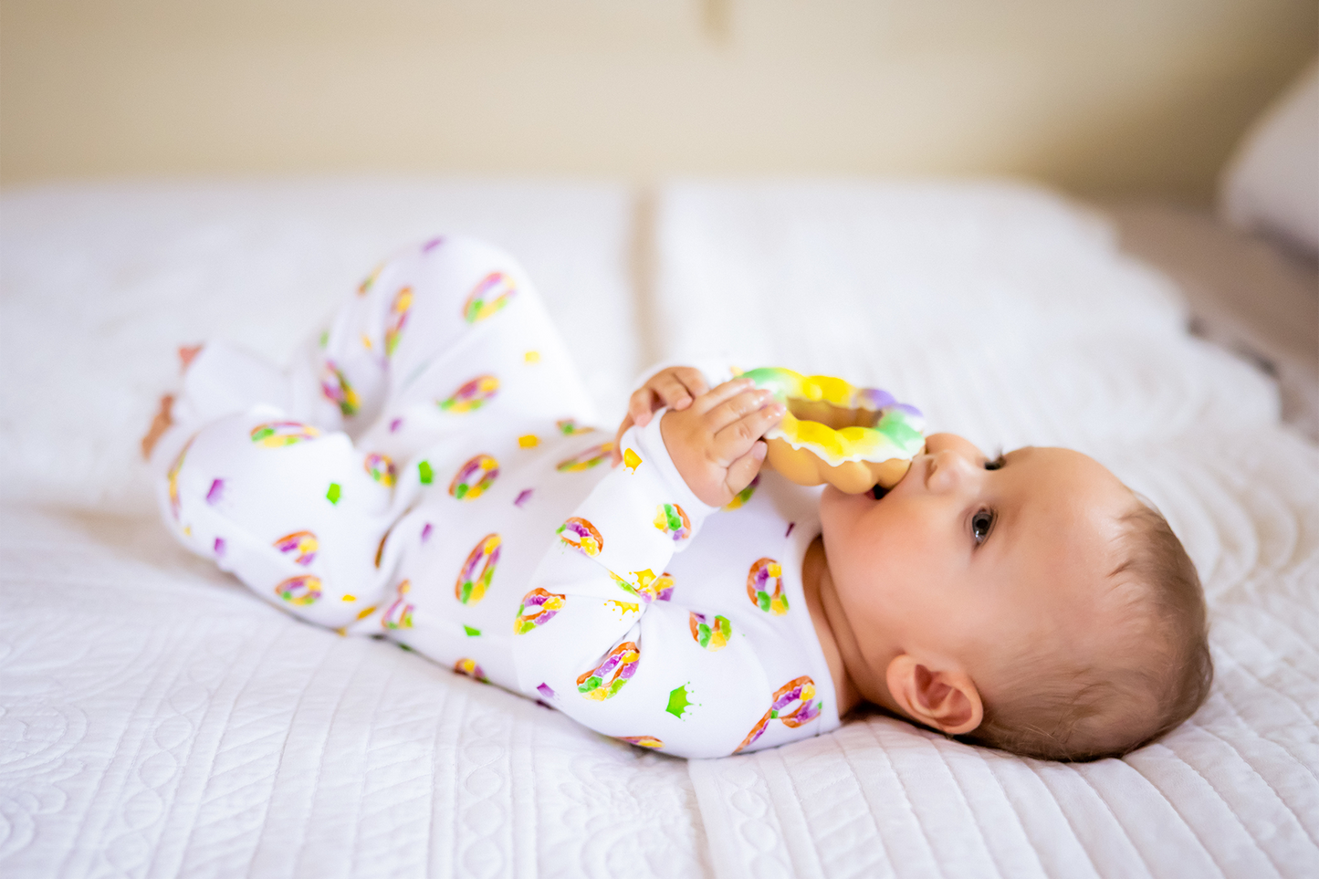 Baby lying on a bed wearing a colorful onesie and holding a toy.