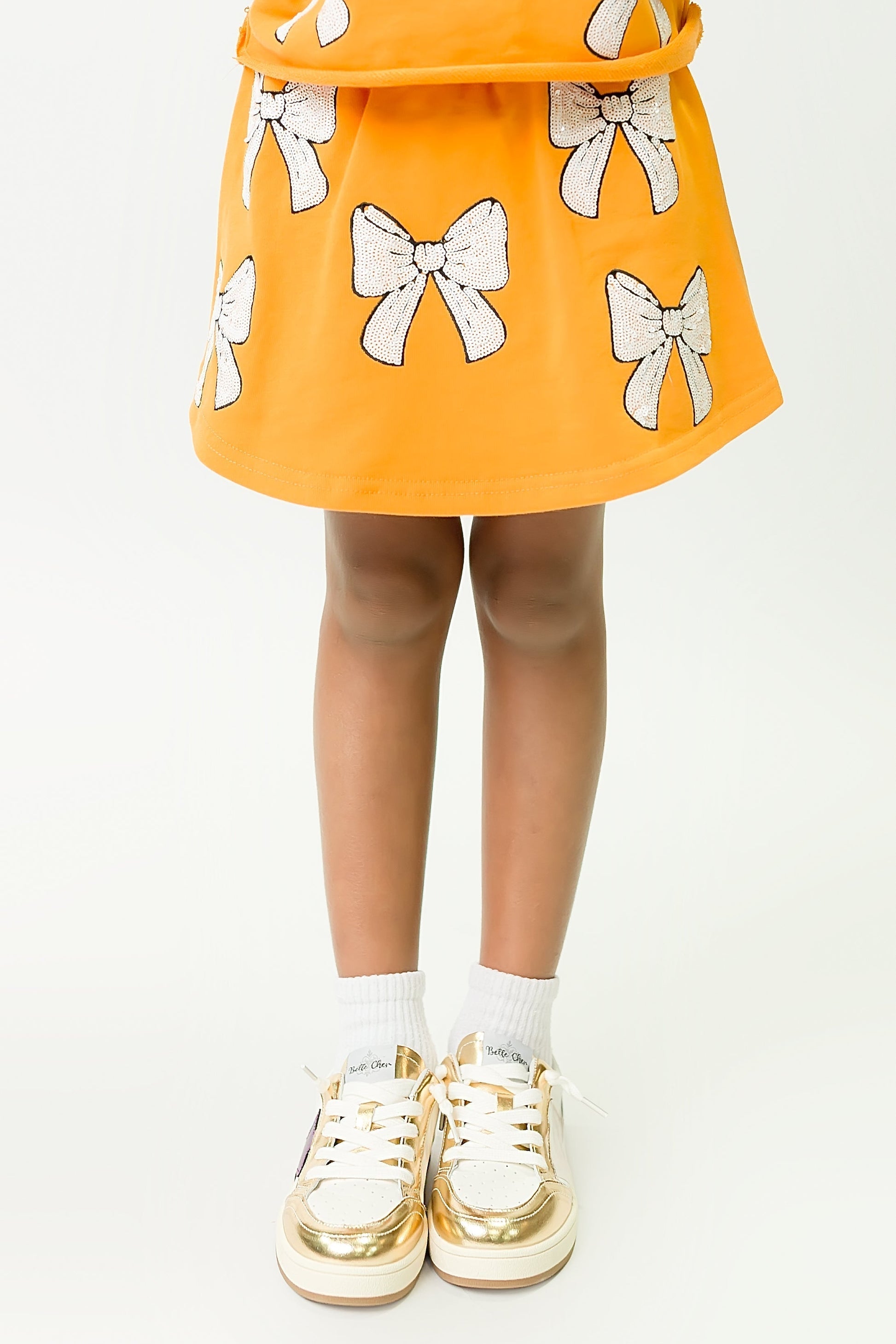Child wearing orange and white bows skort with white bow accents and gold sneakers