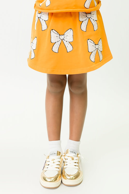 Child wearing orange and white bows skort with white bow accents and gold sneakers