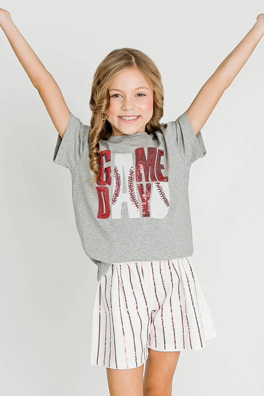 Young girl wearing a gray 'Game Day' t-shirt and white shorts with red stripes, standing against a plain background.