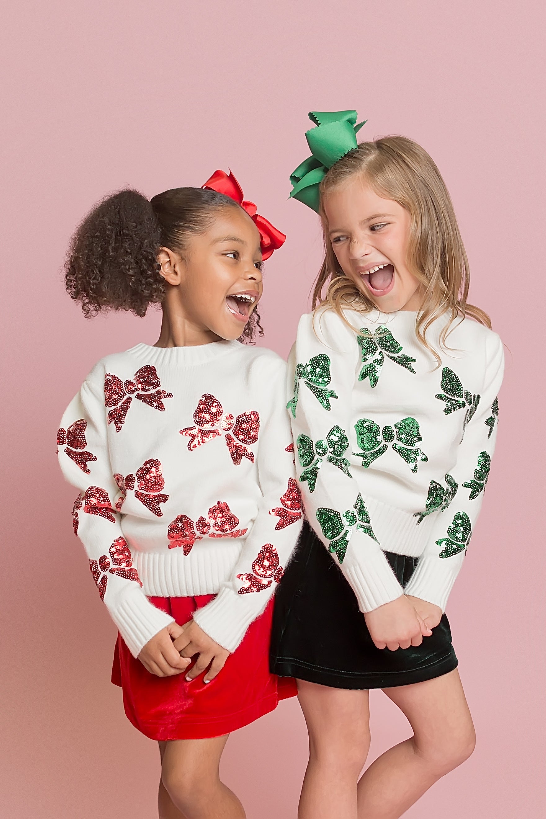 Two children wearing white sweaters with red and green floral patterns on a pink background