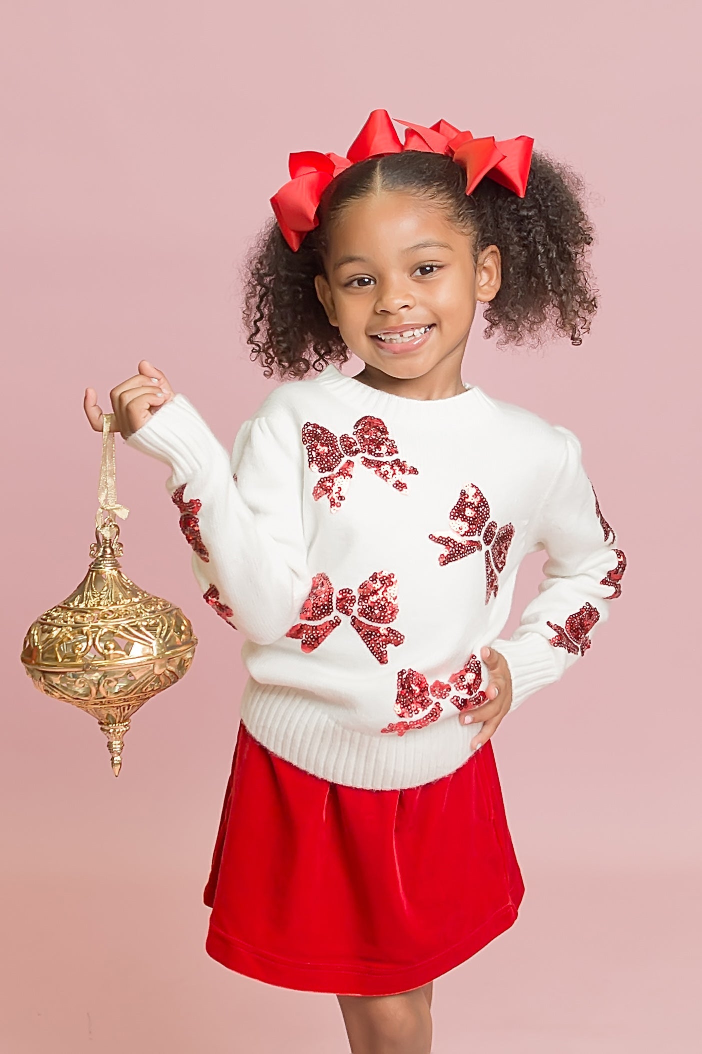 Young girl wearing a white sweater with red floral patterns and a red skirt, holding a gold ornament on a pink background.