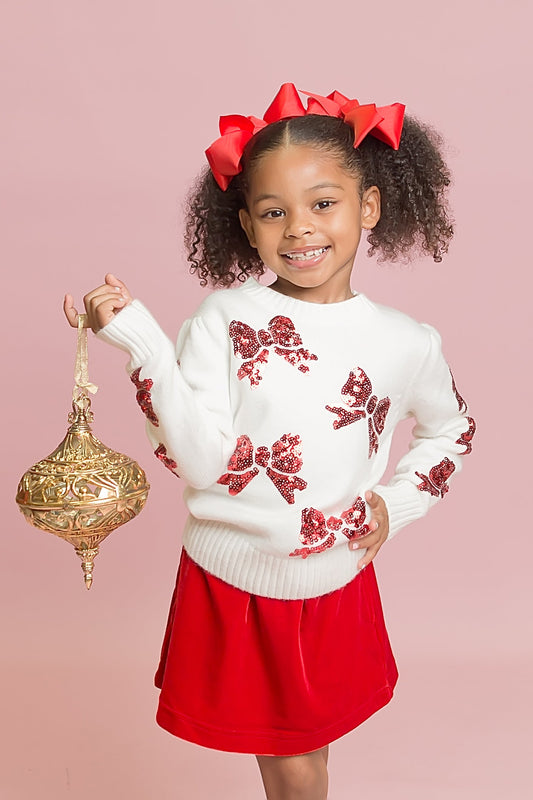 Young girl wearing a white sweater with red floral patterns and a red skirt, holding a gold ornament on a pink background.