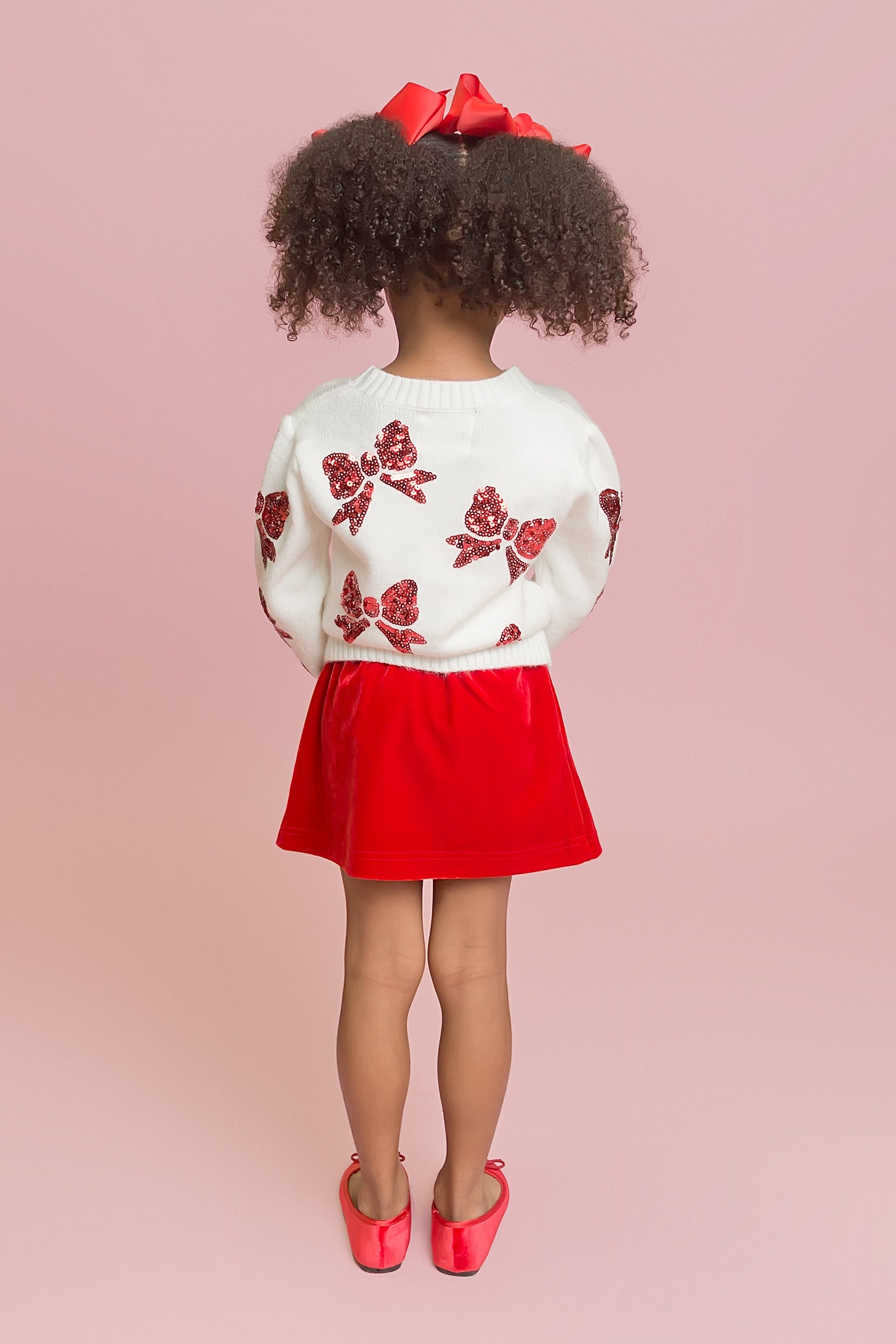 Child wearing a white sweater with red floral patterns and a red skirt on a pink background