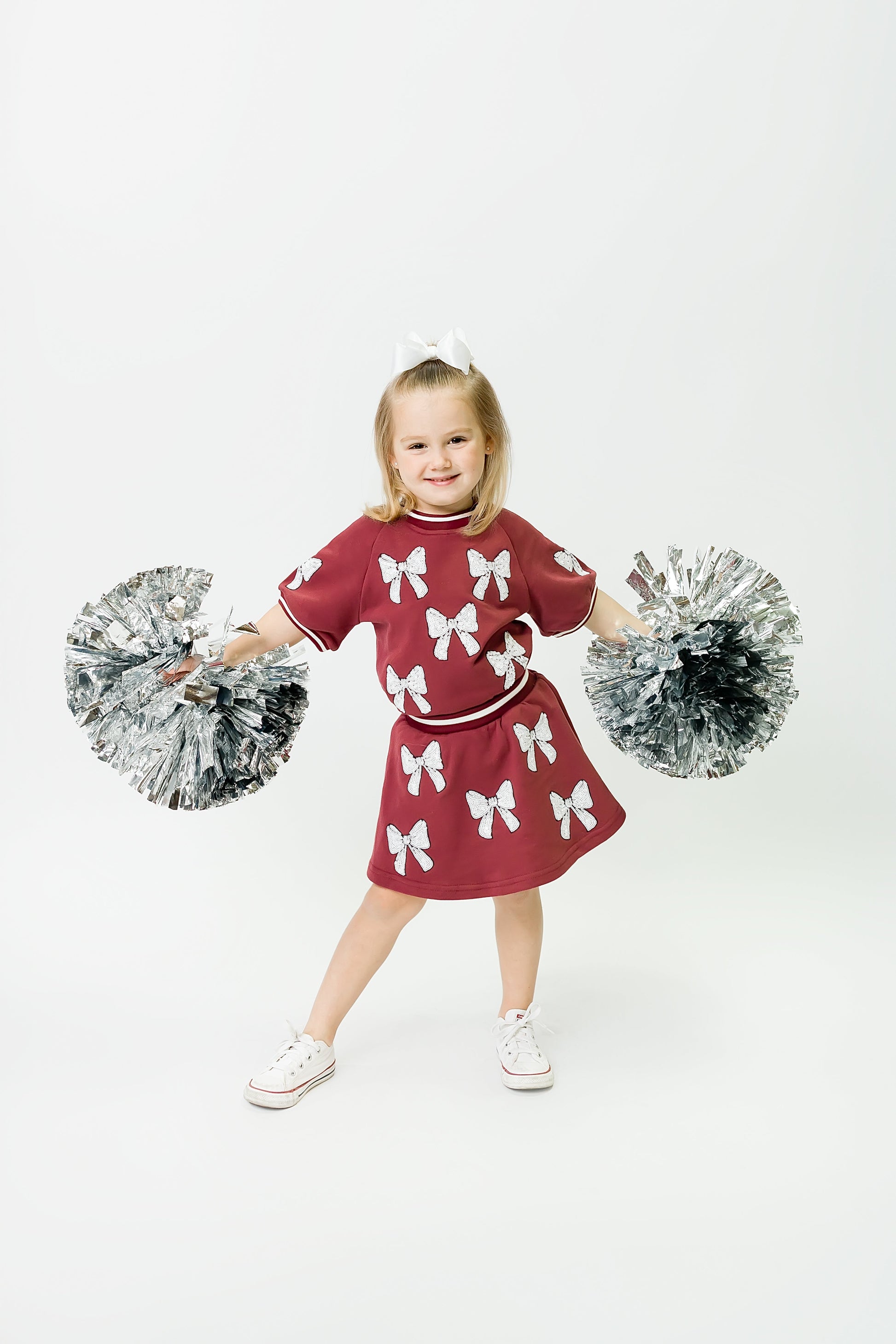 Little girl wearing maroon and white bows skort with pom poms smiling and posing.