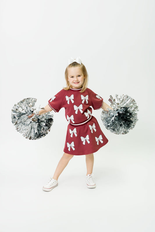 Little girl wearing maroon and white bows skort with pom poms smiling and posing.