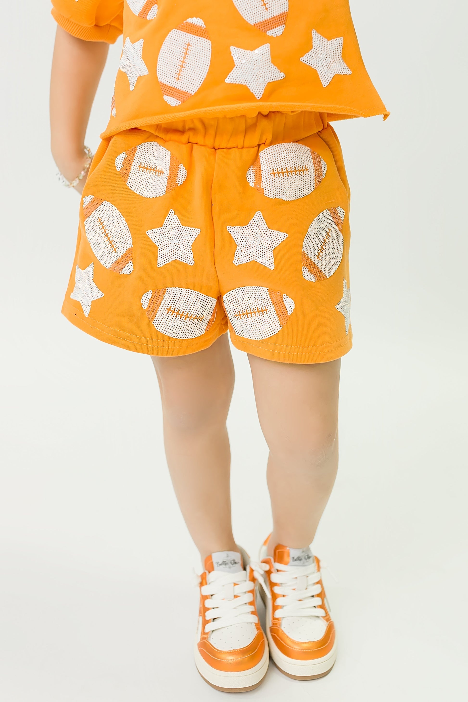 Child wearing orange football shorts with white football and star patterns, paired with matching top and orange sneakers