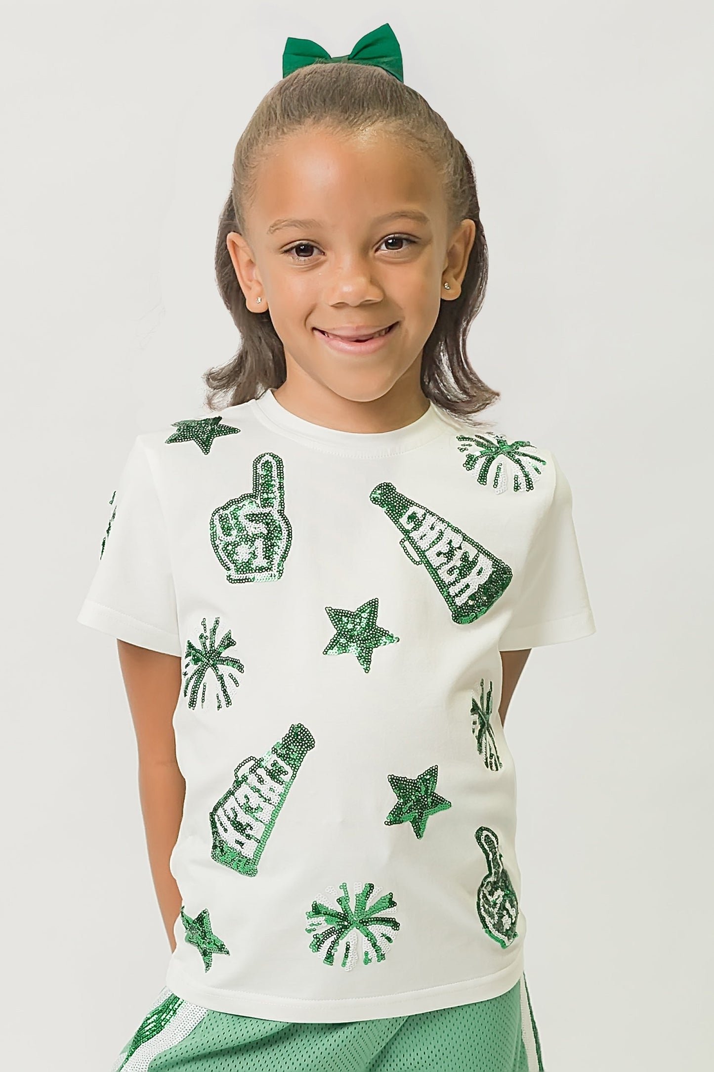 Young girl wearing green and white cheer shirt with sparkling sequin designs and matching green bow