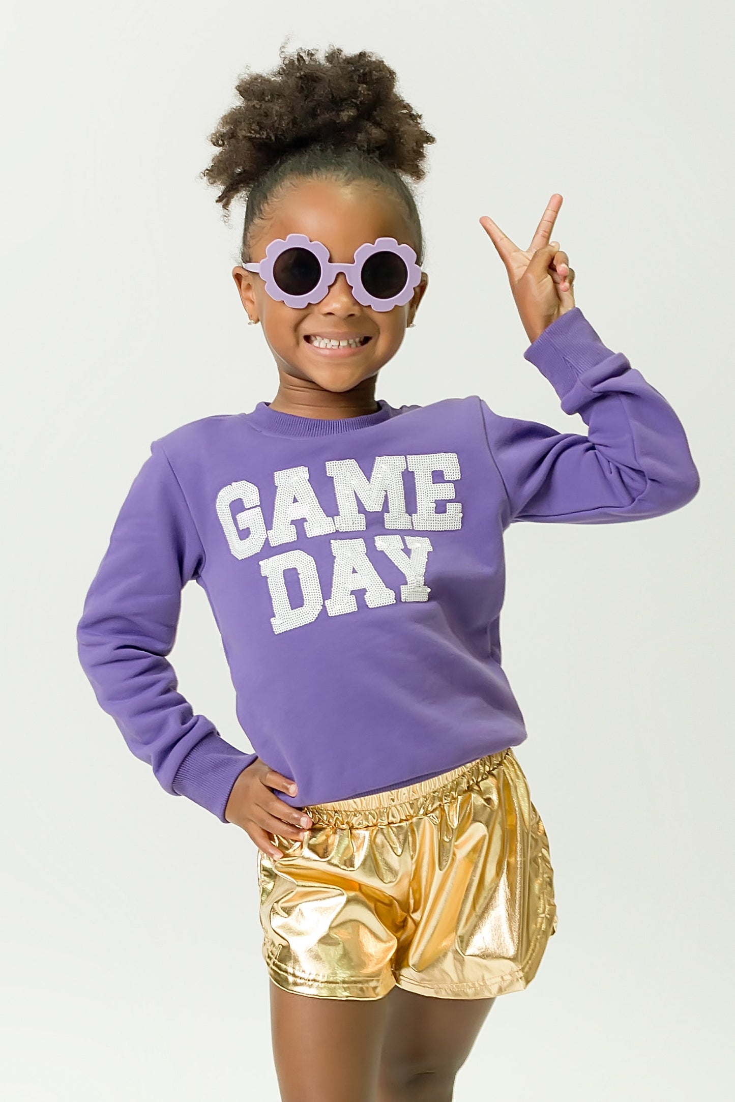 Child wearing purple gameday sweatshirt with "GAME DAY" text and gold shorts smiling with peace sign.