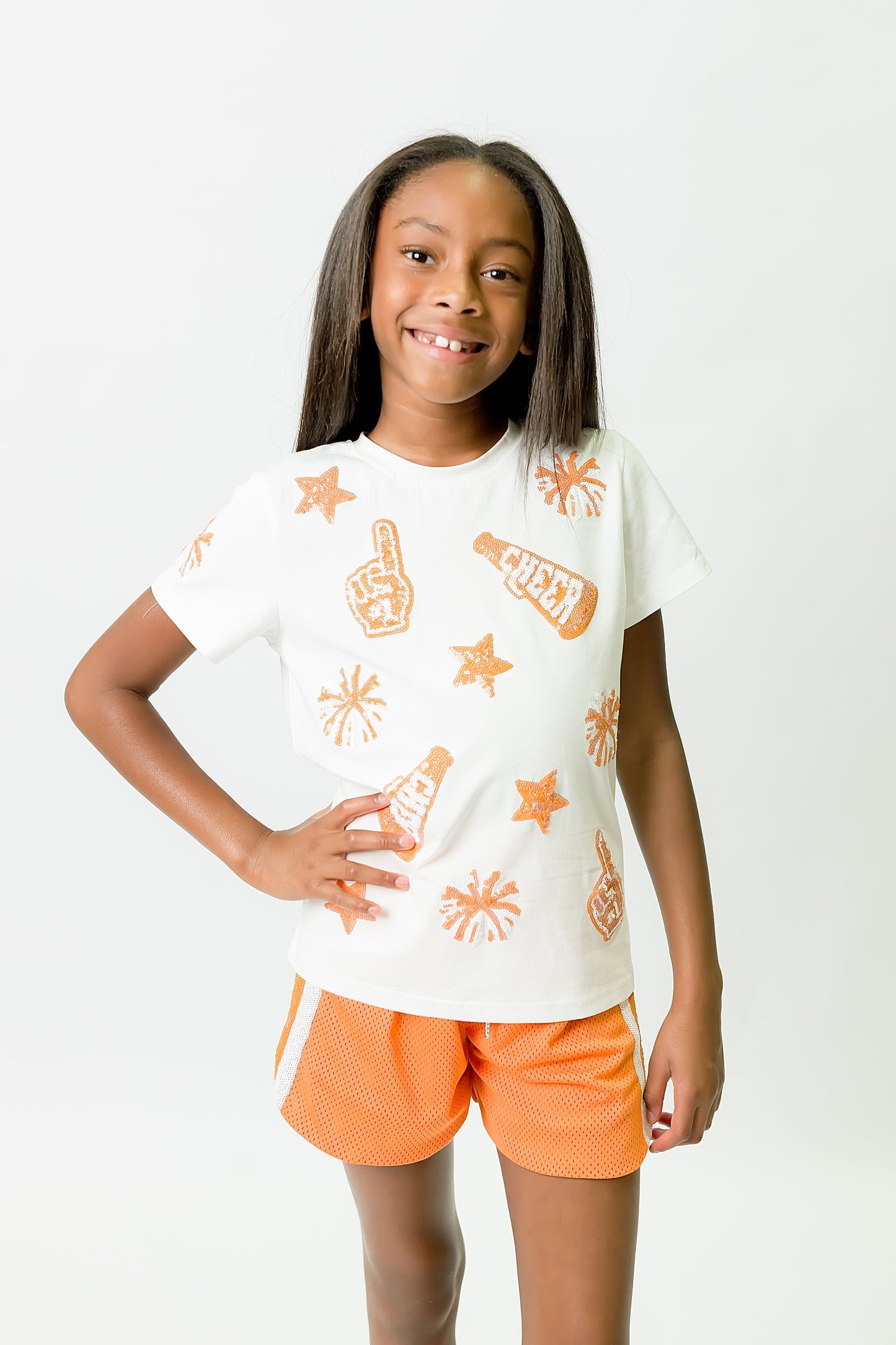 young girl wearing orange and white cheer shirt with sporty orange shorts smiling confidently
