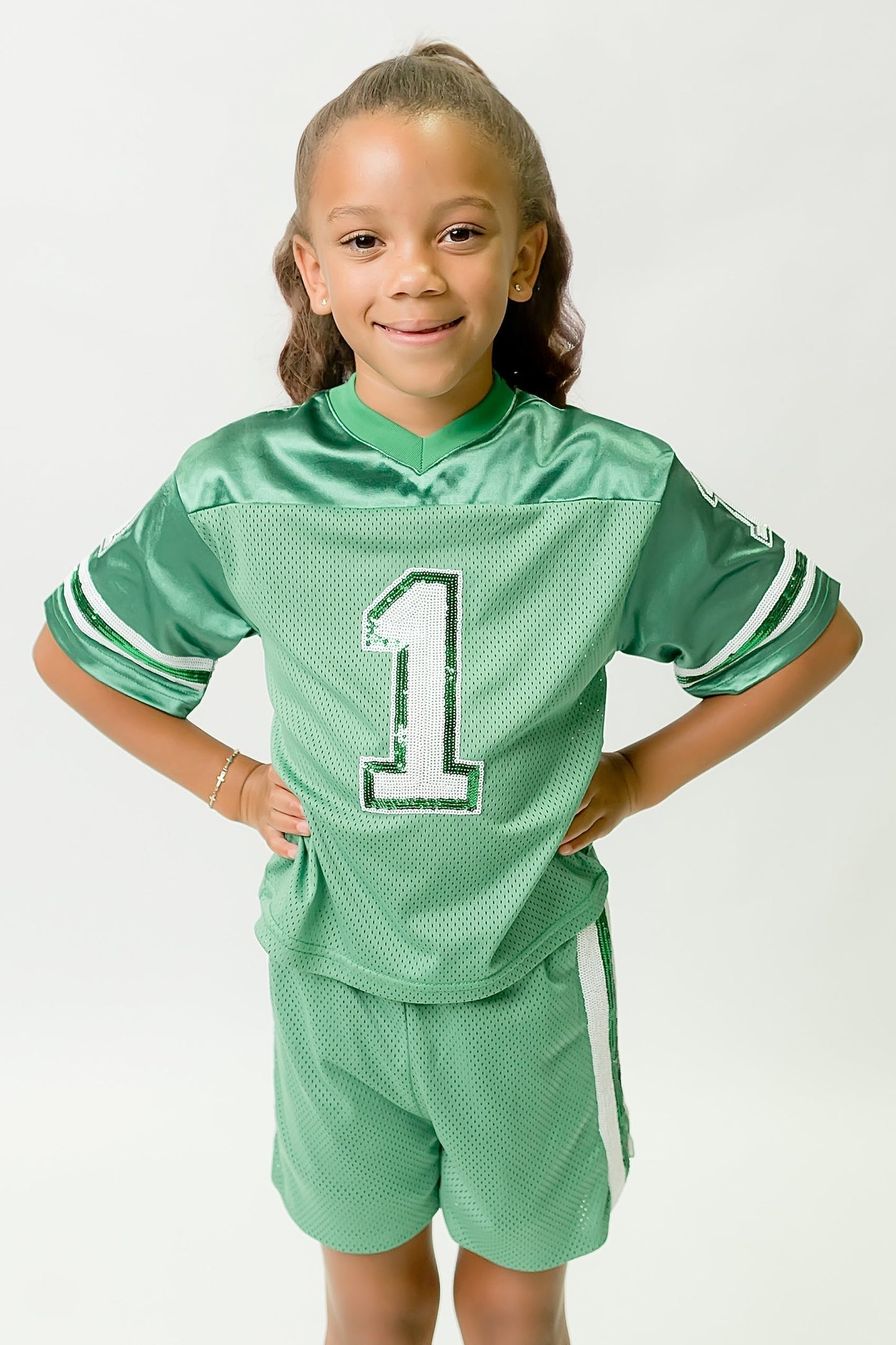 Young girl wearing green and white sequin jersey with number 1 standing with hands on hips