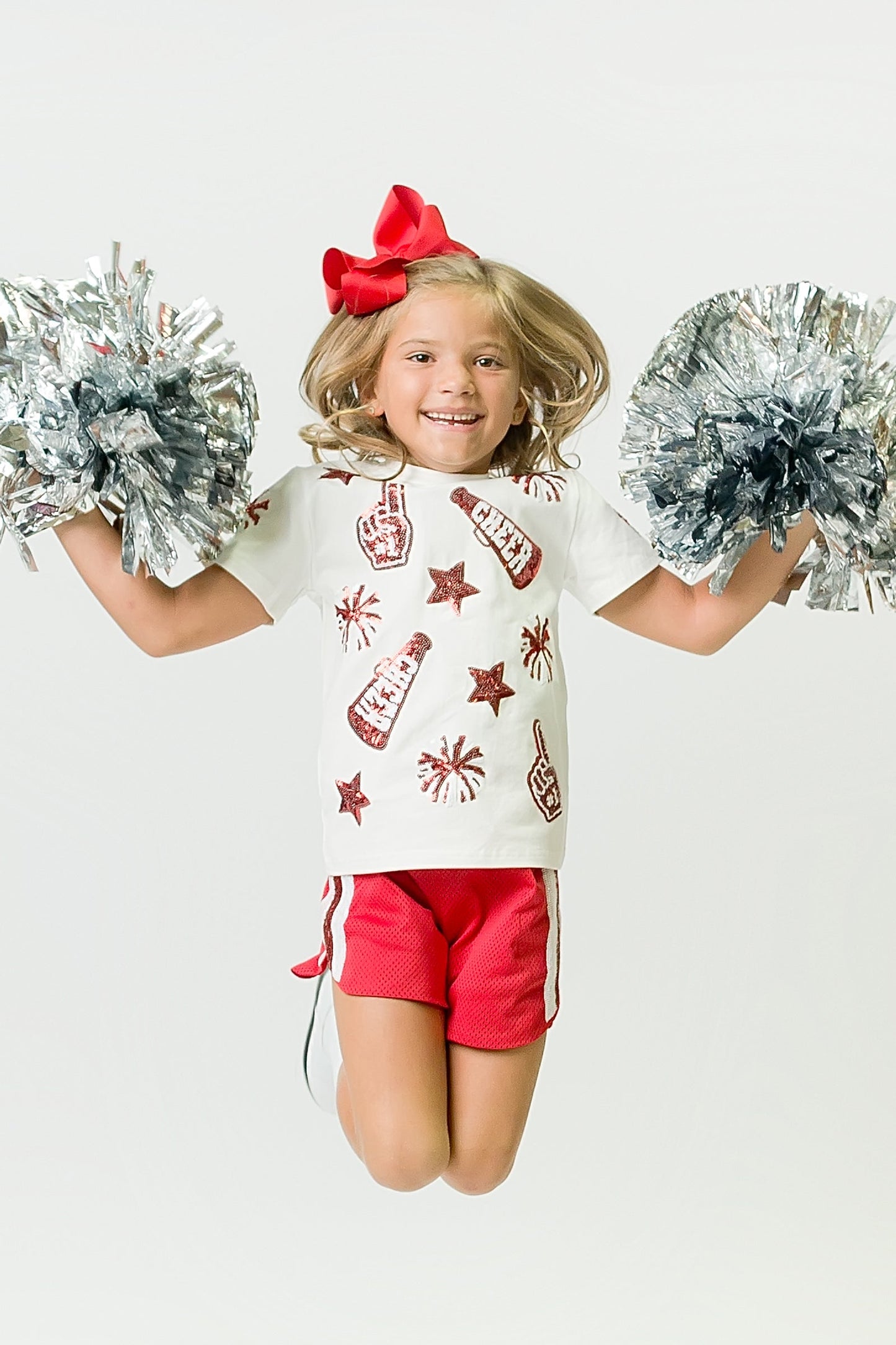Young girl jumping wearing a red and white cheer shirt with red sequins and holding silver pom-poms