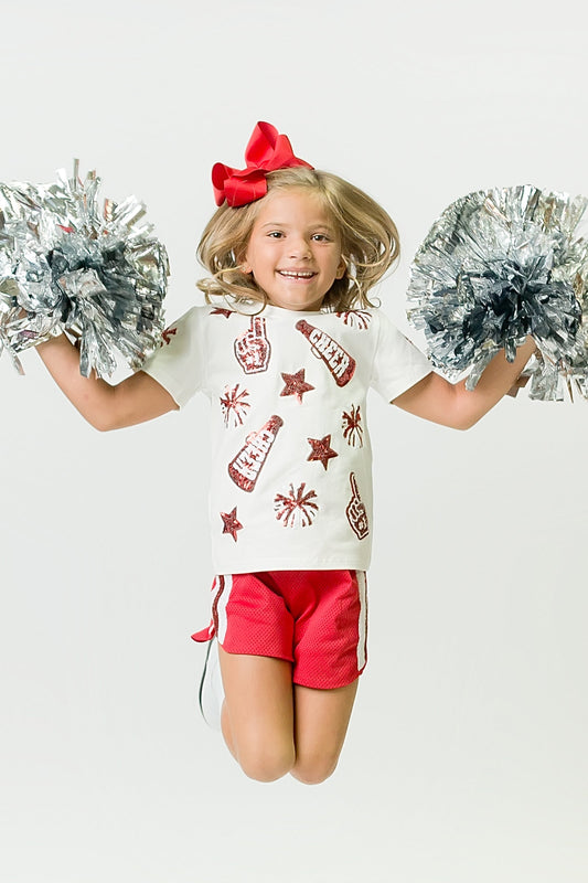 Young girl jumping wearing a red and white cheer shirt with red sequins and holding silver pom-poms