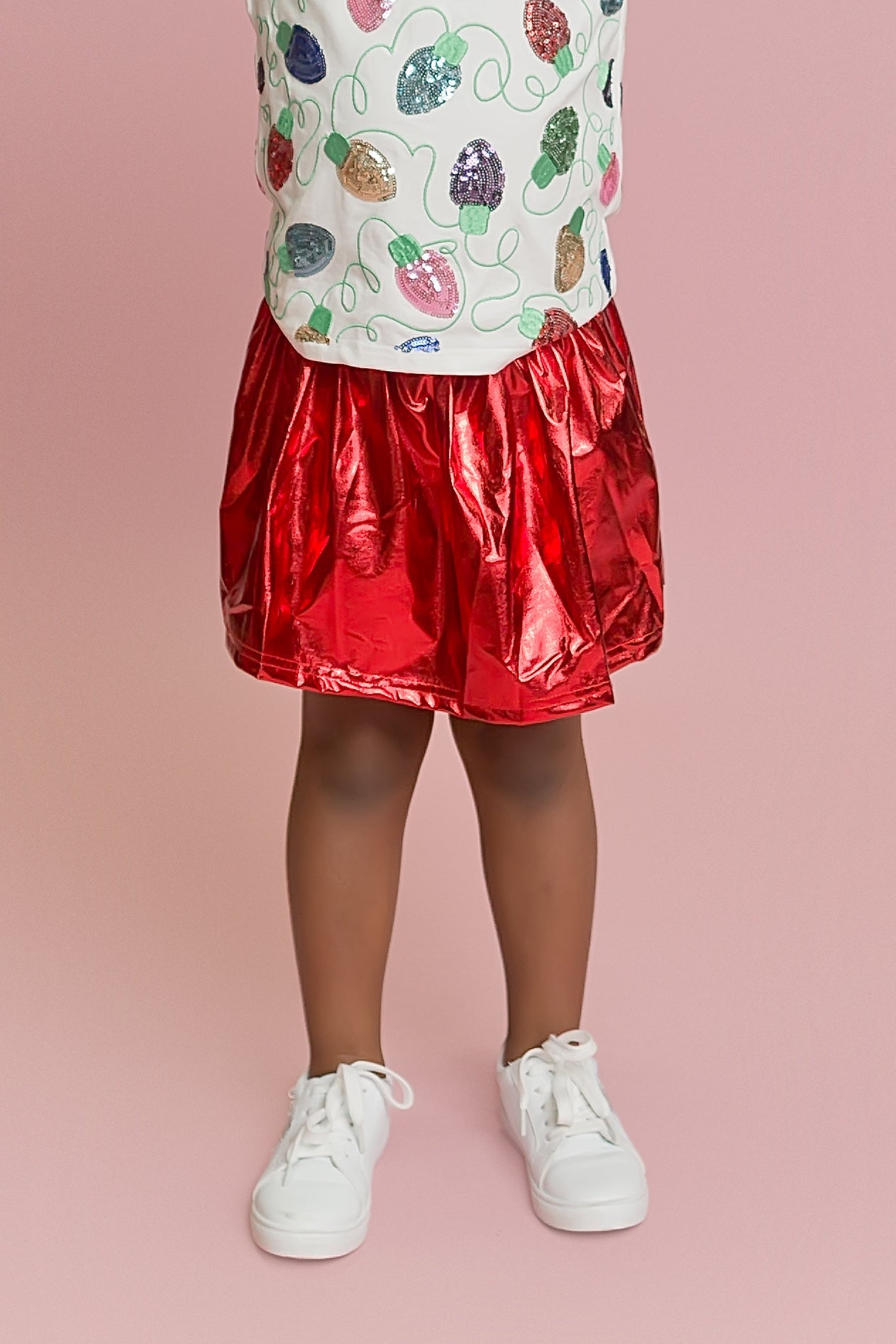 Child wearing a colorful patterned top and red metallic skirt on a pink background