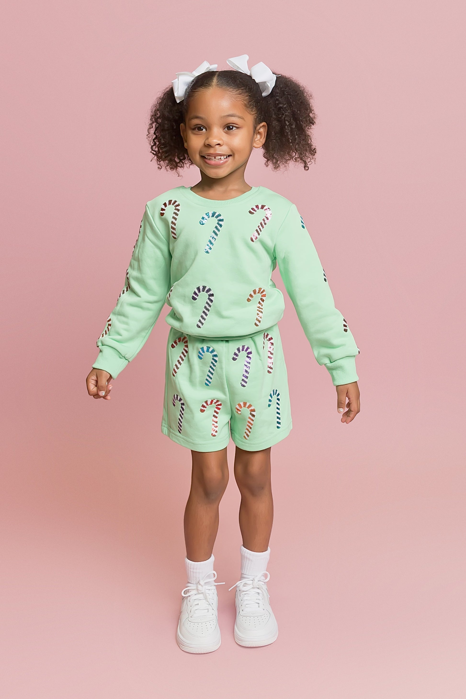 Young girl wearing a matching green sweater and shorts set with candy cane designs on a pink background