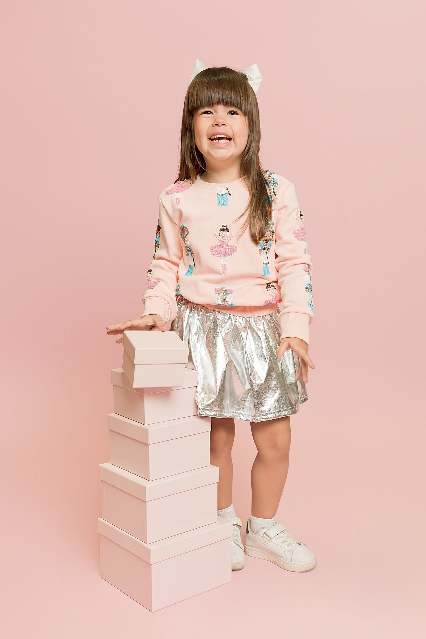 Child in pink sweater and silver skirt standing next to stacked blocks on a pink background