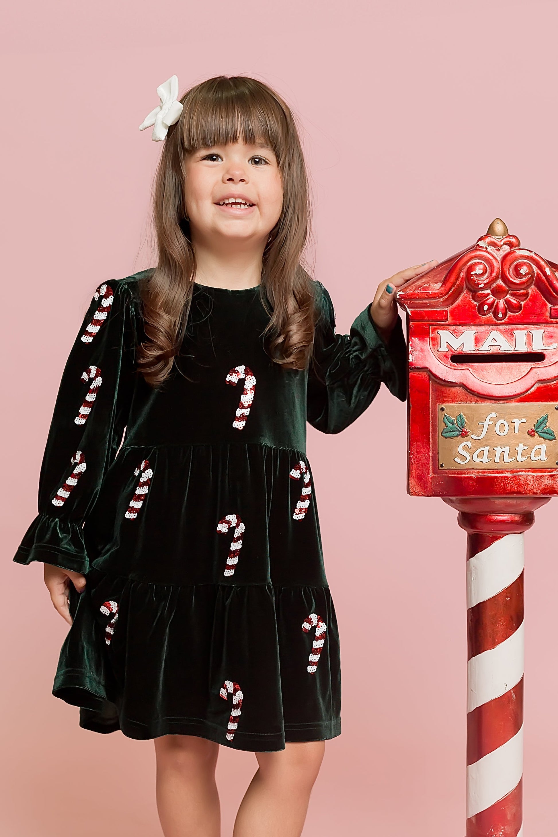 Young girl in a festive outfit standing next to a red mailbox with candy cane design on a pink background