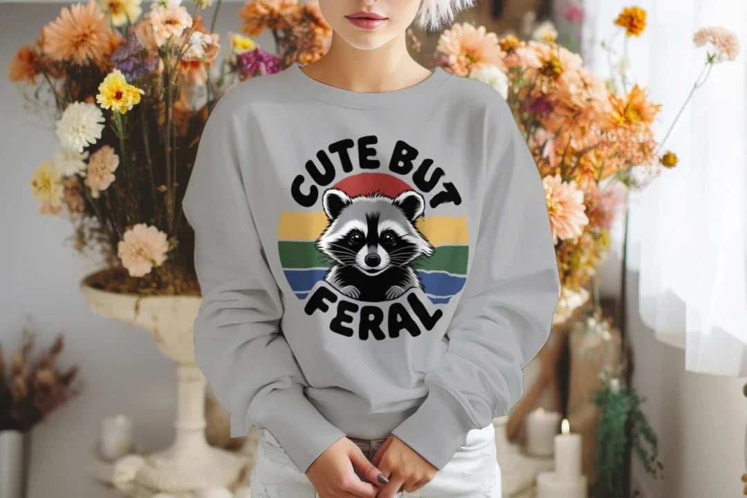 Person wearing a gray sweatshirt with a raccoon graphic and text 'Cute But Feral' in a decorated indoor setting.