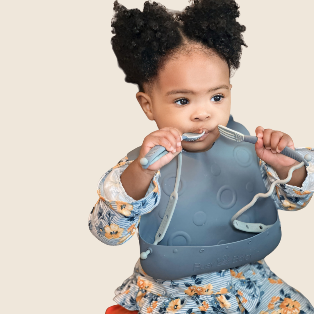 baby wearing a gray Travel Tether Bib with attached utensils holding a spoon and fork