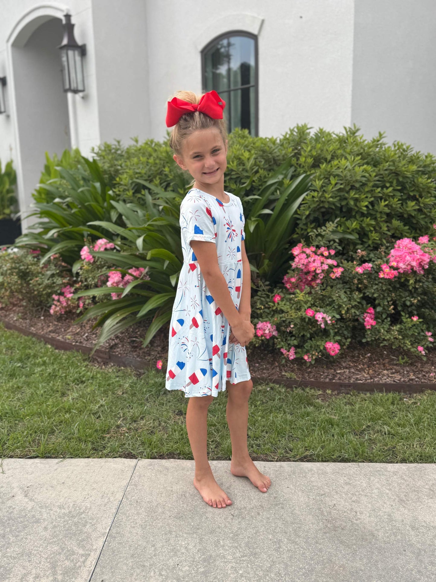 Girl wearing a Popsicles and Fireworks Play Dress standing outside with greenery and pink flowers.