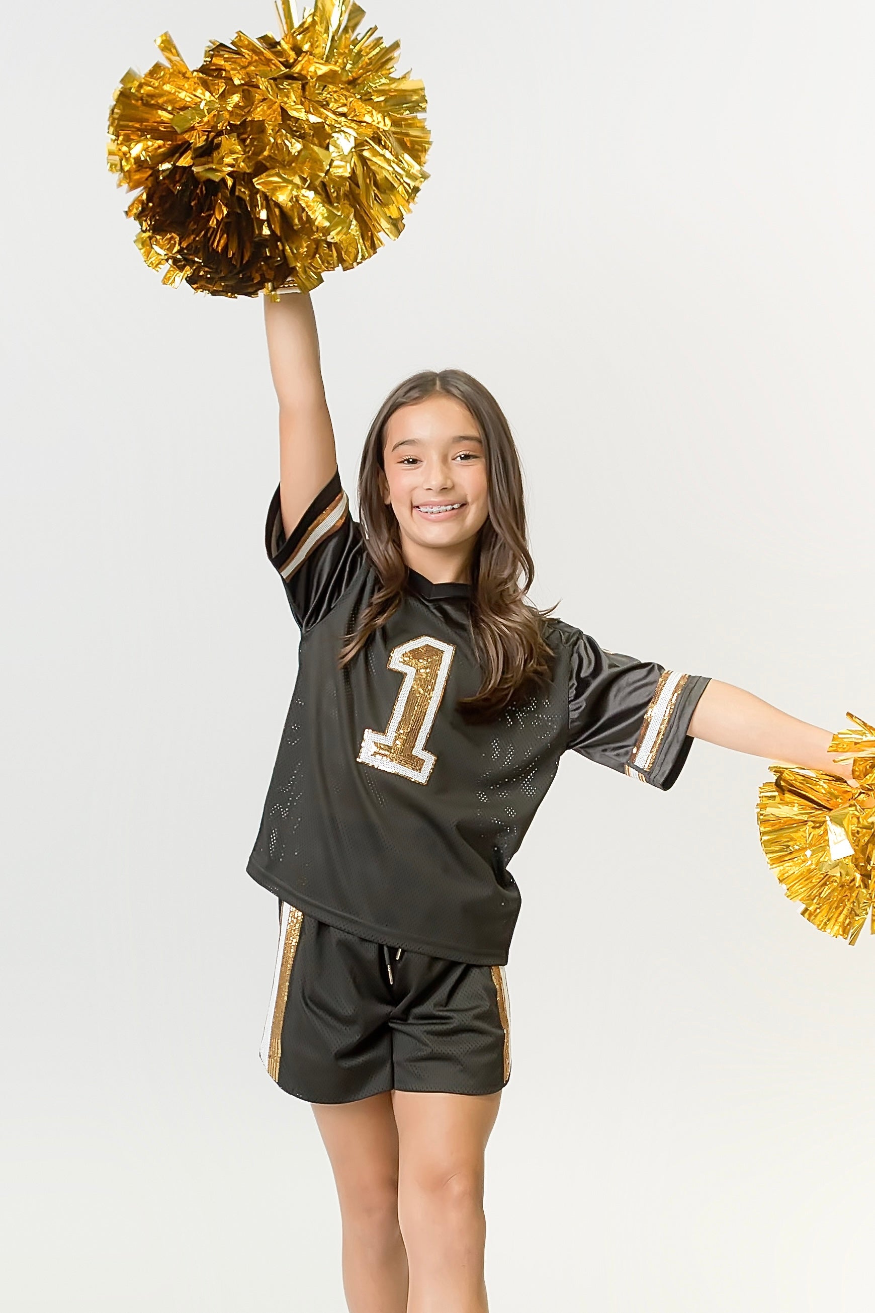 Young cheerleader wearing black and gold sequin jersey with number 1 and matching shorts holding gold pom-poms