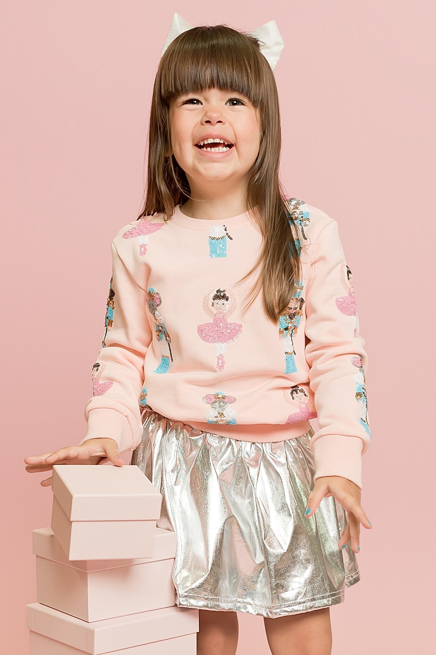 Child wearing a pink sweater with cupcake design and a silver skirt, standing against a pink background.