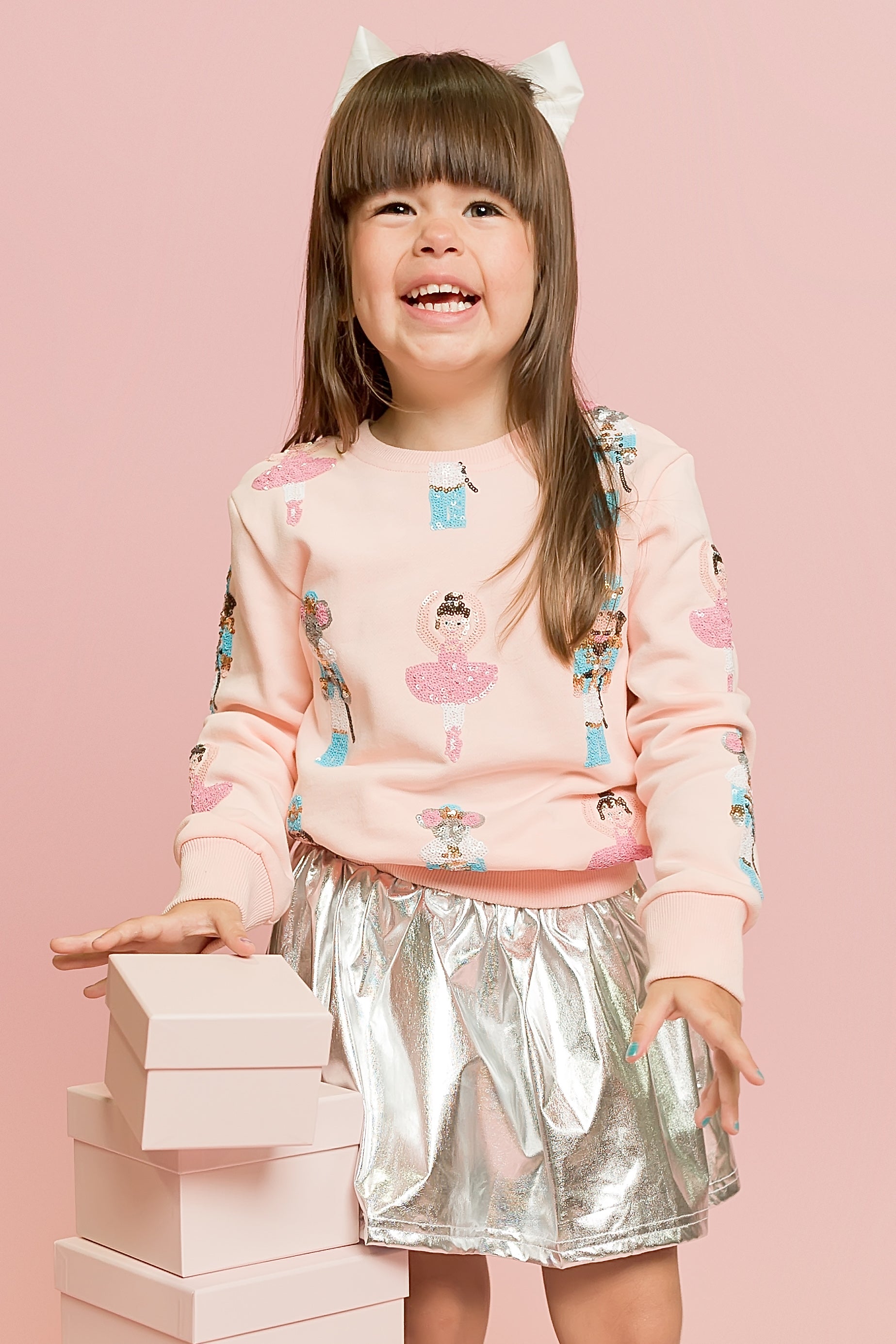 Child wearing a pink sweater with cupcake design and a silver skirt, standing against a pink background.