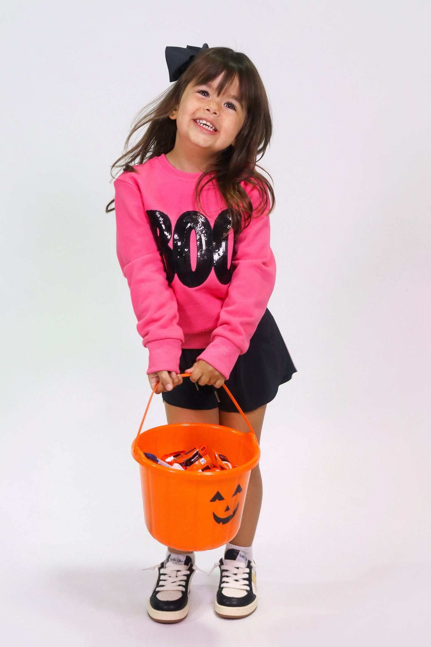 Girl wearing hot pink boo sweatshirt with black sequins holding orange Halloween candy bucket