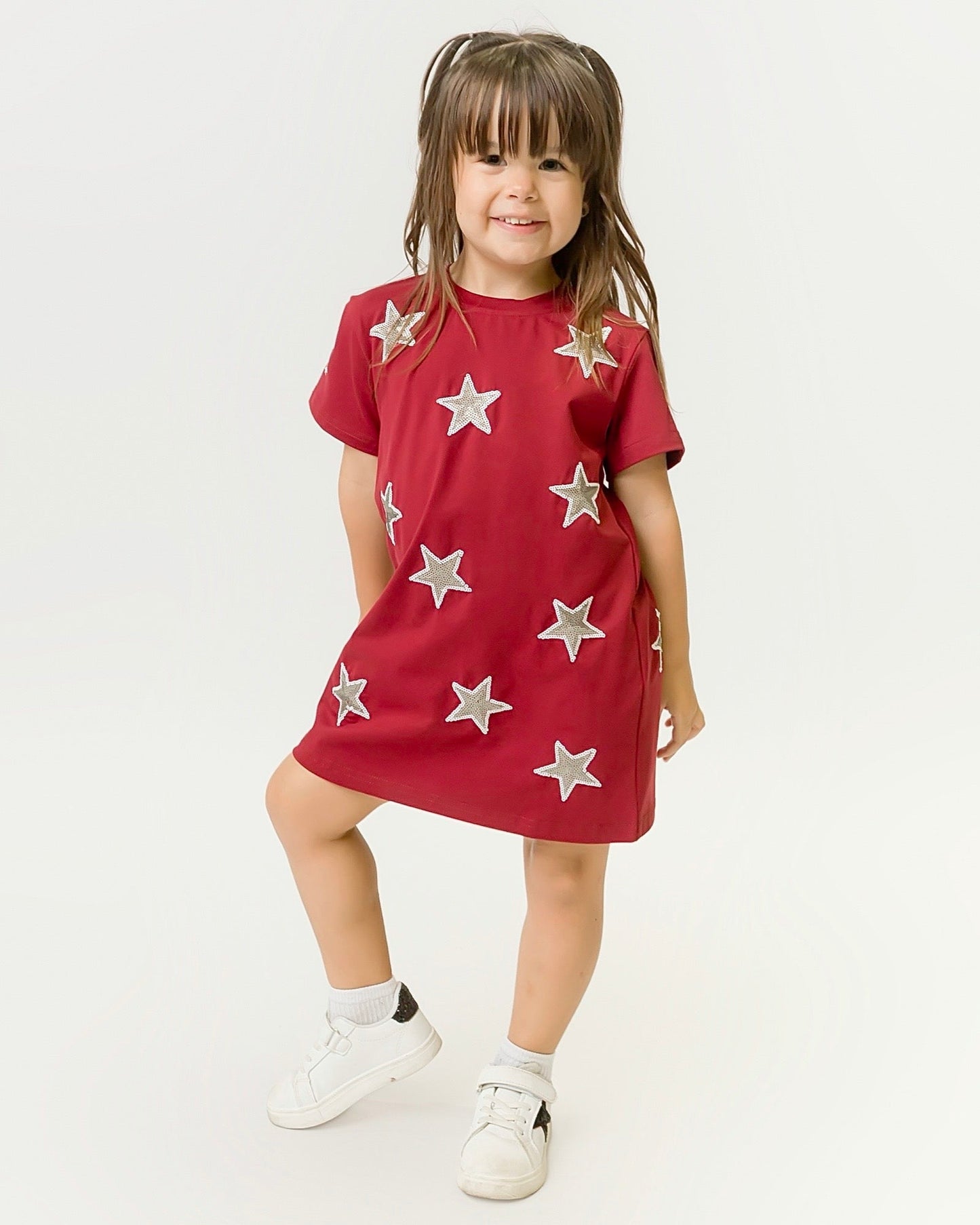 Little girl wearing maroon and gray star kids dress with white sneakers smiling and posing against a plain background