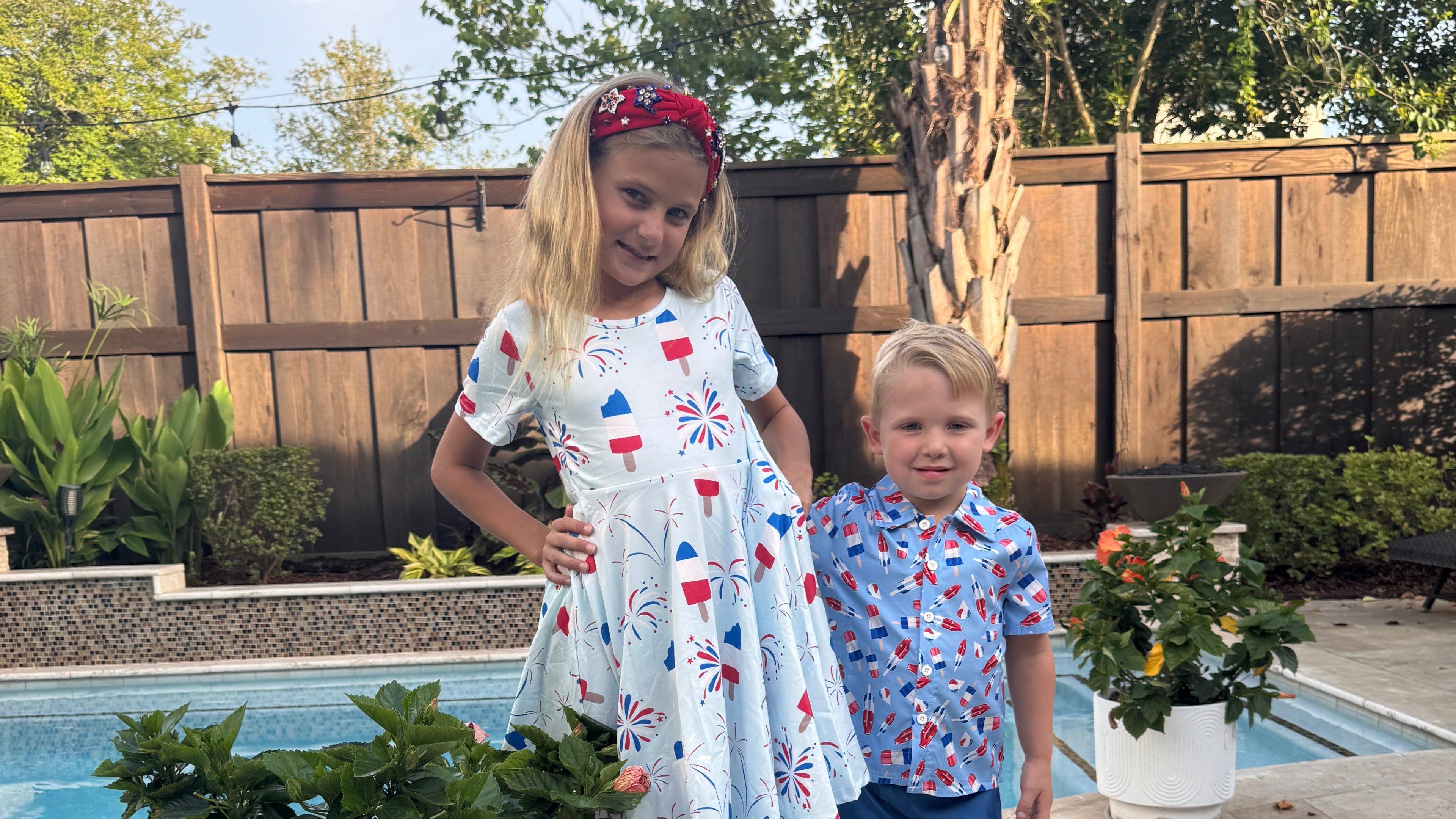 Two children standing by a pool with plants and a wooden deck in the background