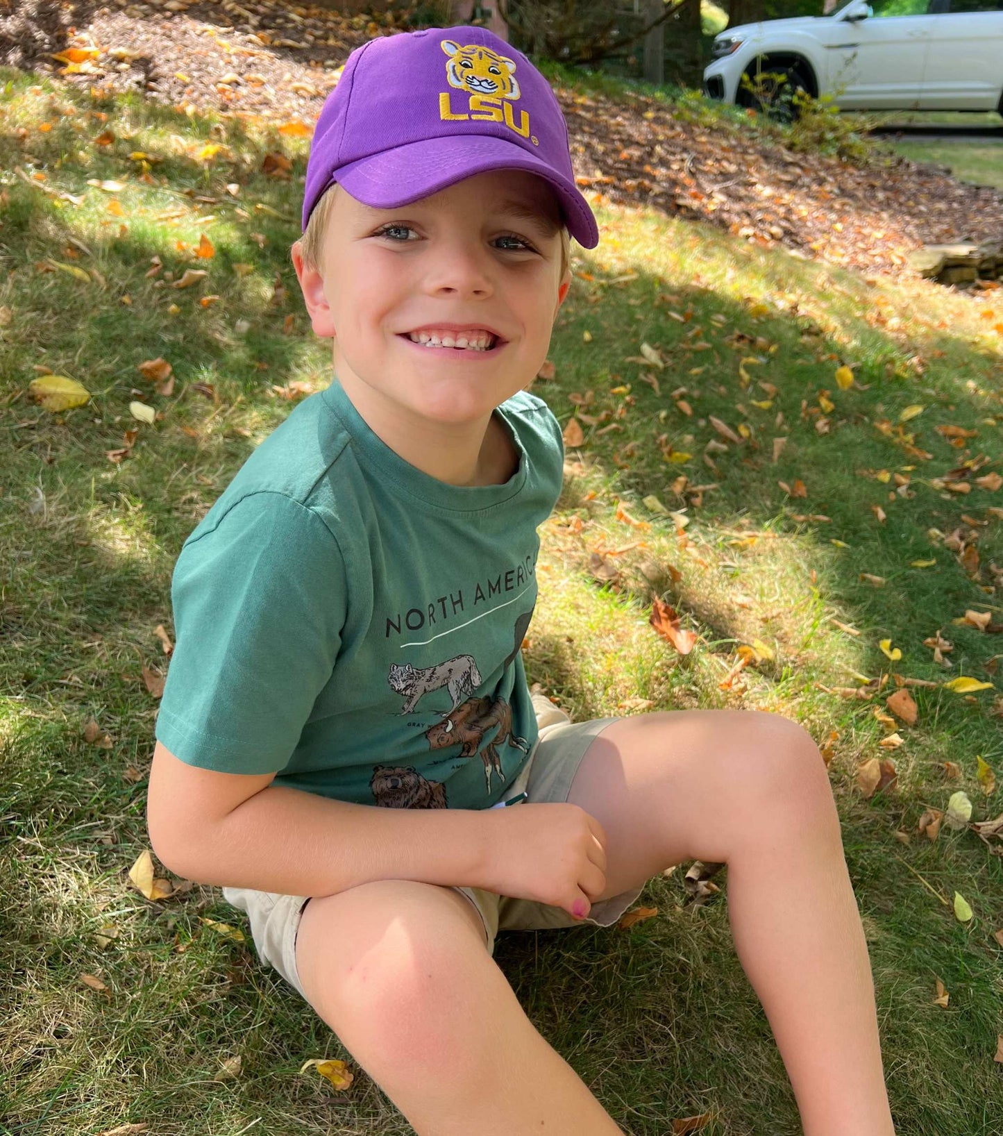 Child wearing a purple LSU cap sitting on grass with fallen leaves