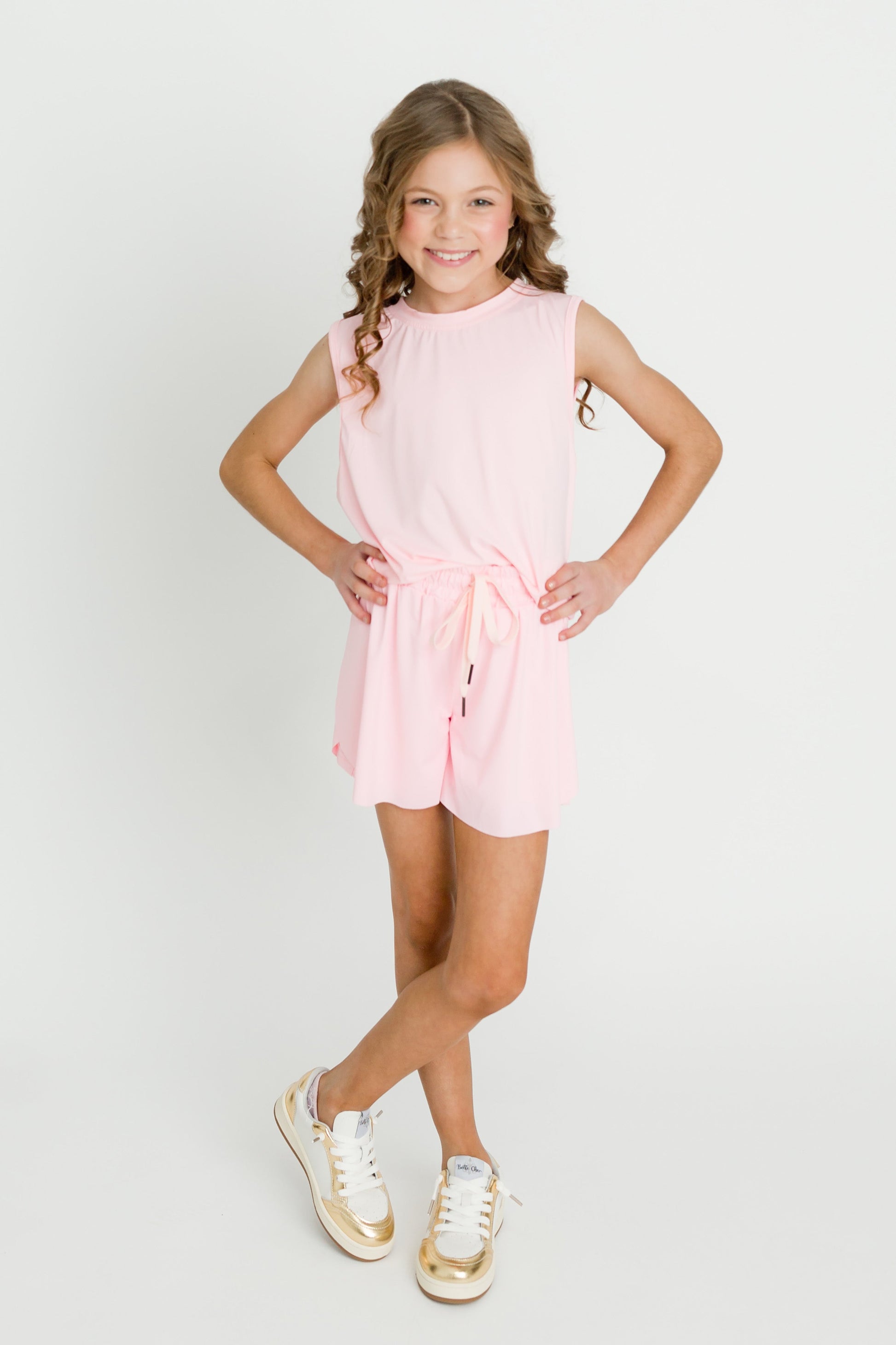 Little girl wearing light pink high low kid tank with tied waist and white gold sneakers