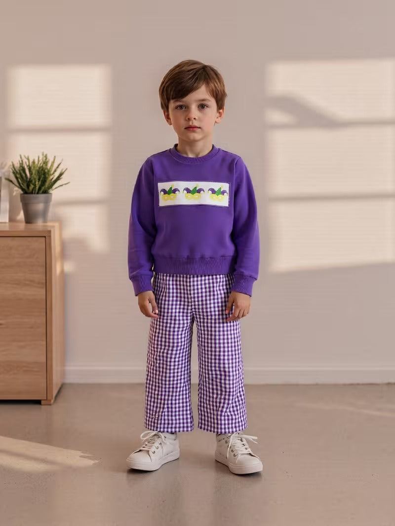 Child wearing a purple sweater with a graphic and checkered pants in a room with a wooden cabinet and plant.