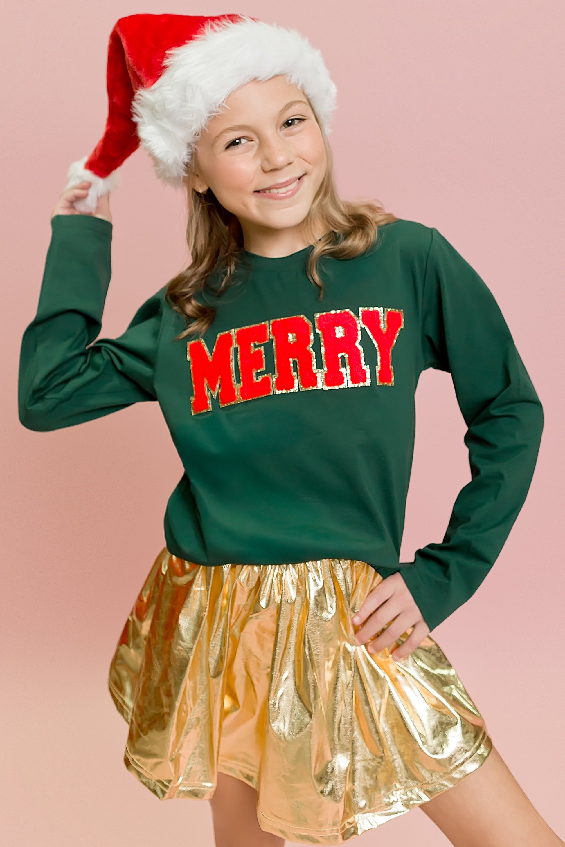 Child wearing a green sweater with 'MERRY' in red, gold skirt, and Santa hat on a pink background