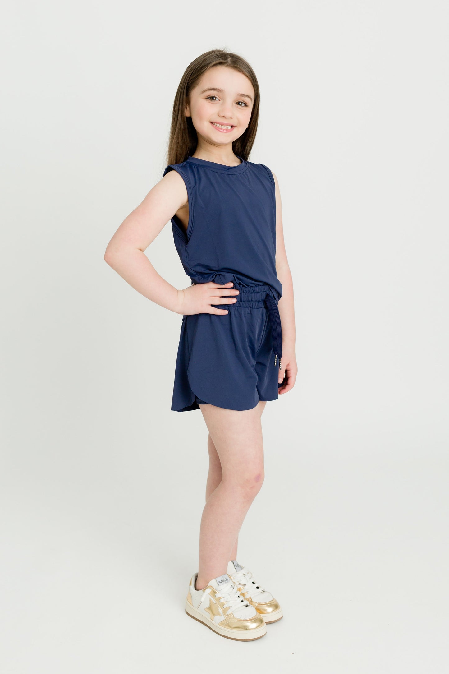 Young girl wearing navy blue high low kid tank and matching shorts with gold sneakers