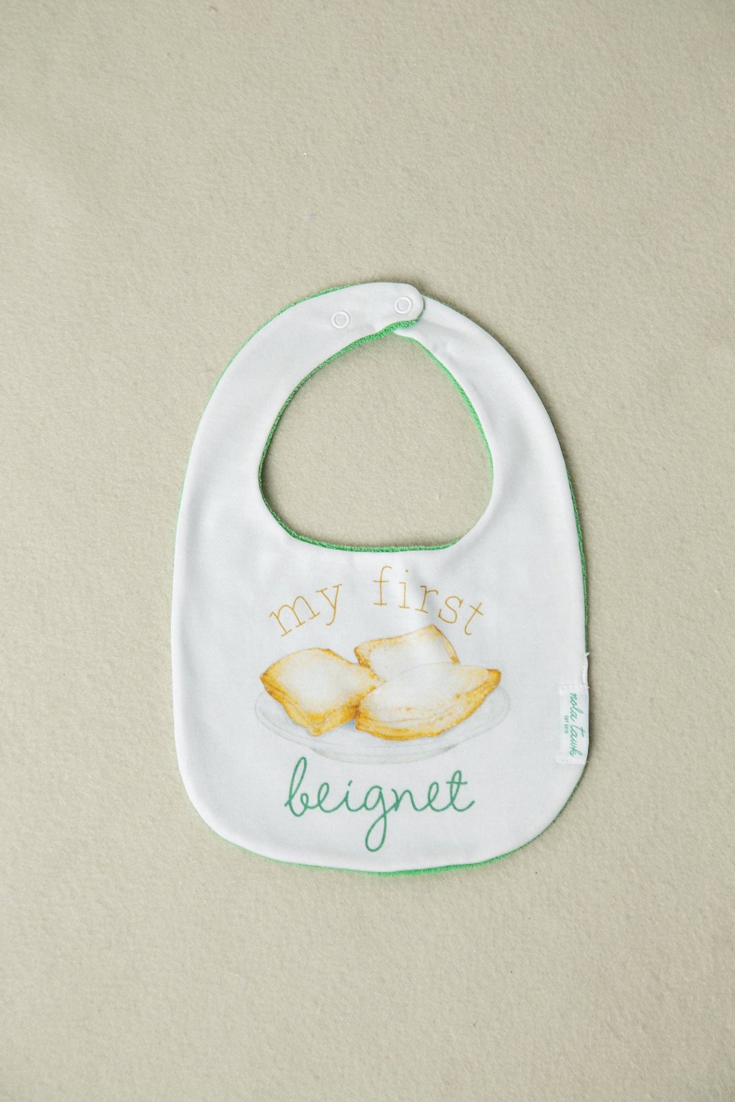 Baby bib with text and beignet illustration on a beige background