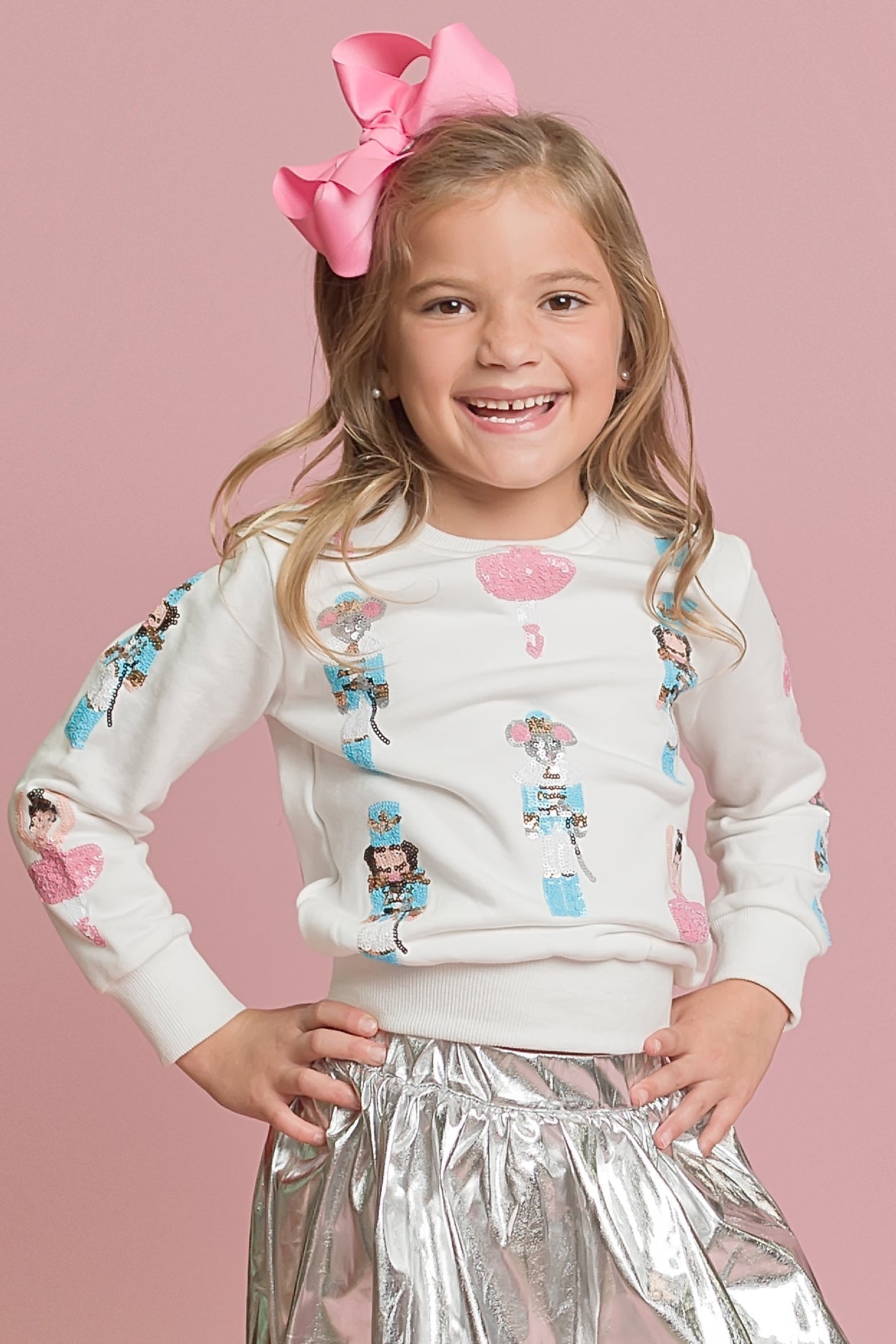 Young girl wearing a white sweater with cartoon characters and a pink bow on her head against a pink background