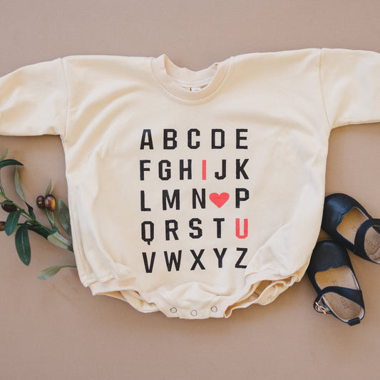 Baby onesie with alphabet print on a beige surface with shoes and leaves.