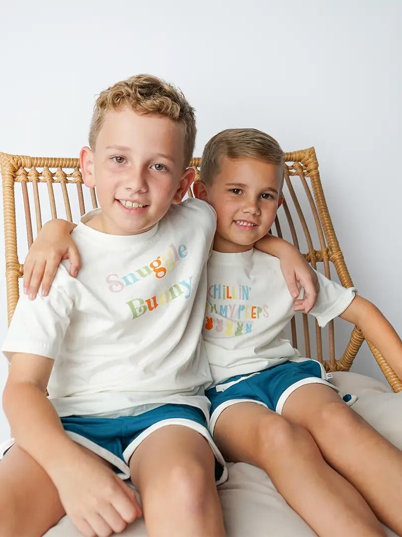 Toddler boys wearing cream Easter cotton tee shirts with colorful graphics, sitting on a rattan chair.